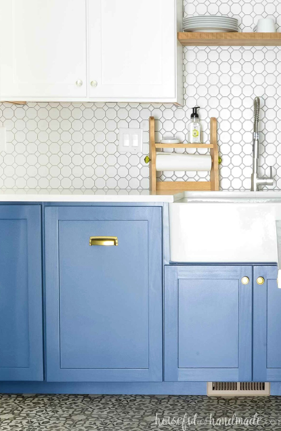 Blue and white kitchen featuring trash cans concealed behind a cabinet door beside a white farmhouse sink.