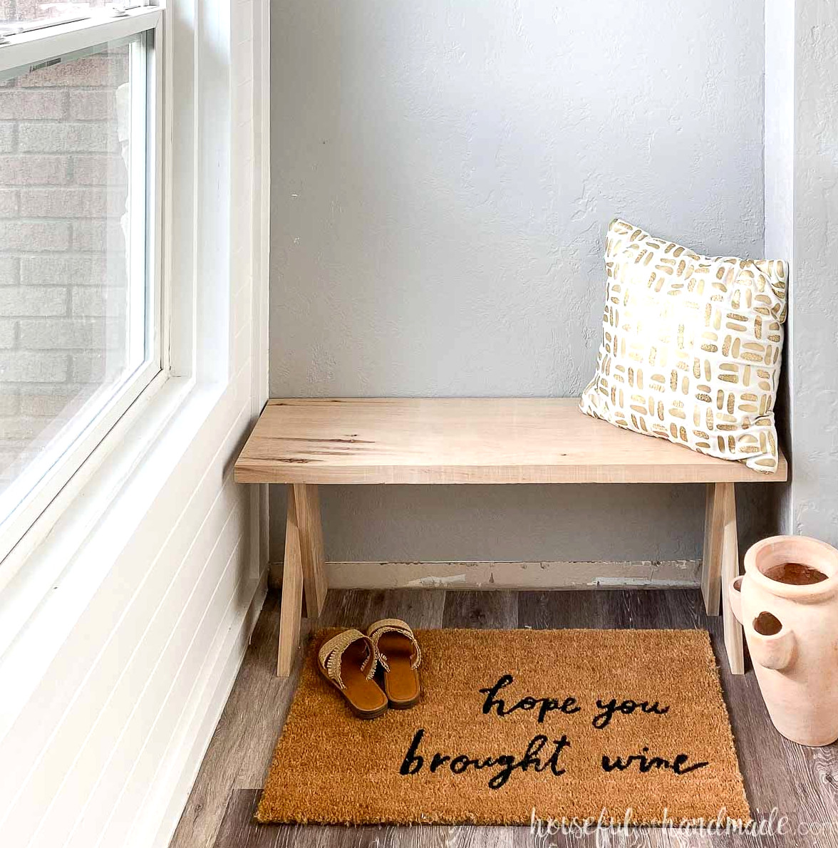 DIY x-leg bench made from hickory wood in an entryway with driftwood vinyl flooring and "hope you brought wine" rug in front with accessories.