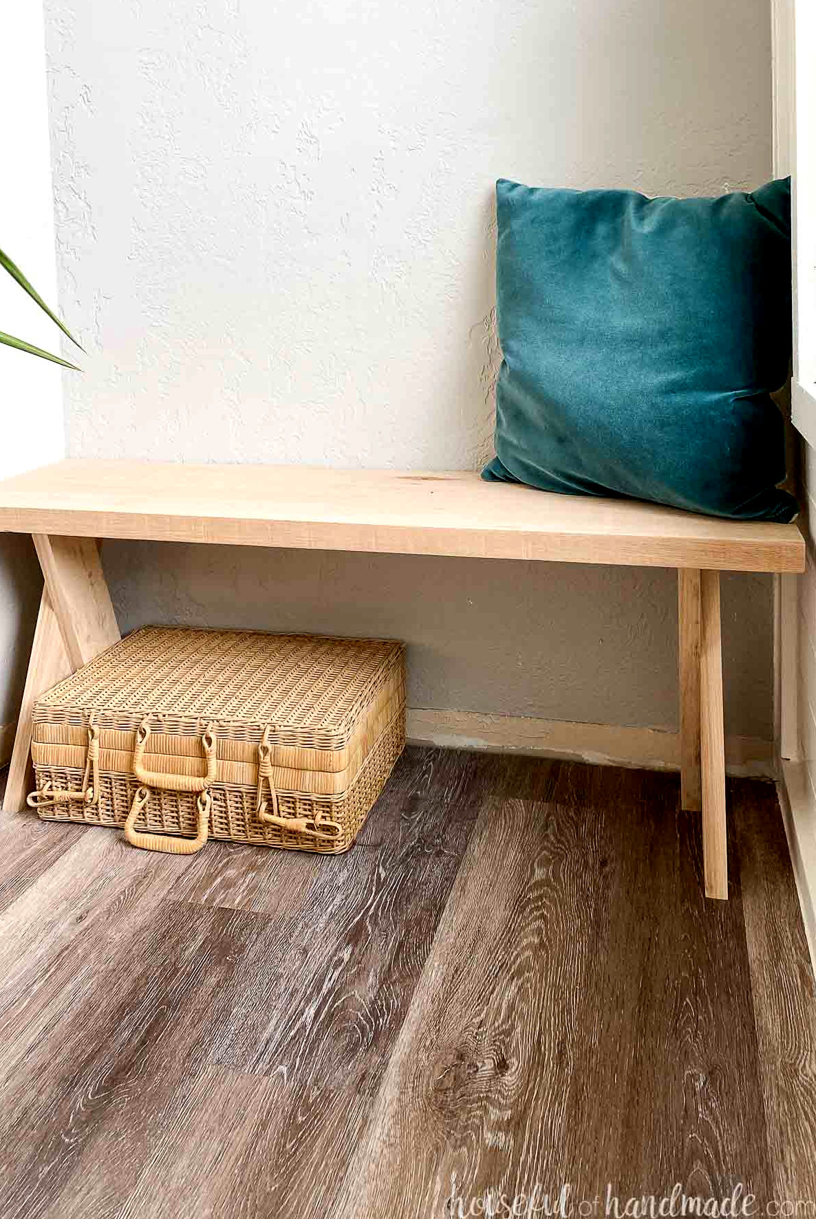 Close view of the DIY bench x-legs with a blue velvet pillow above and a wicker suitcase beneath.