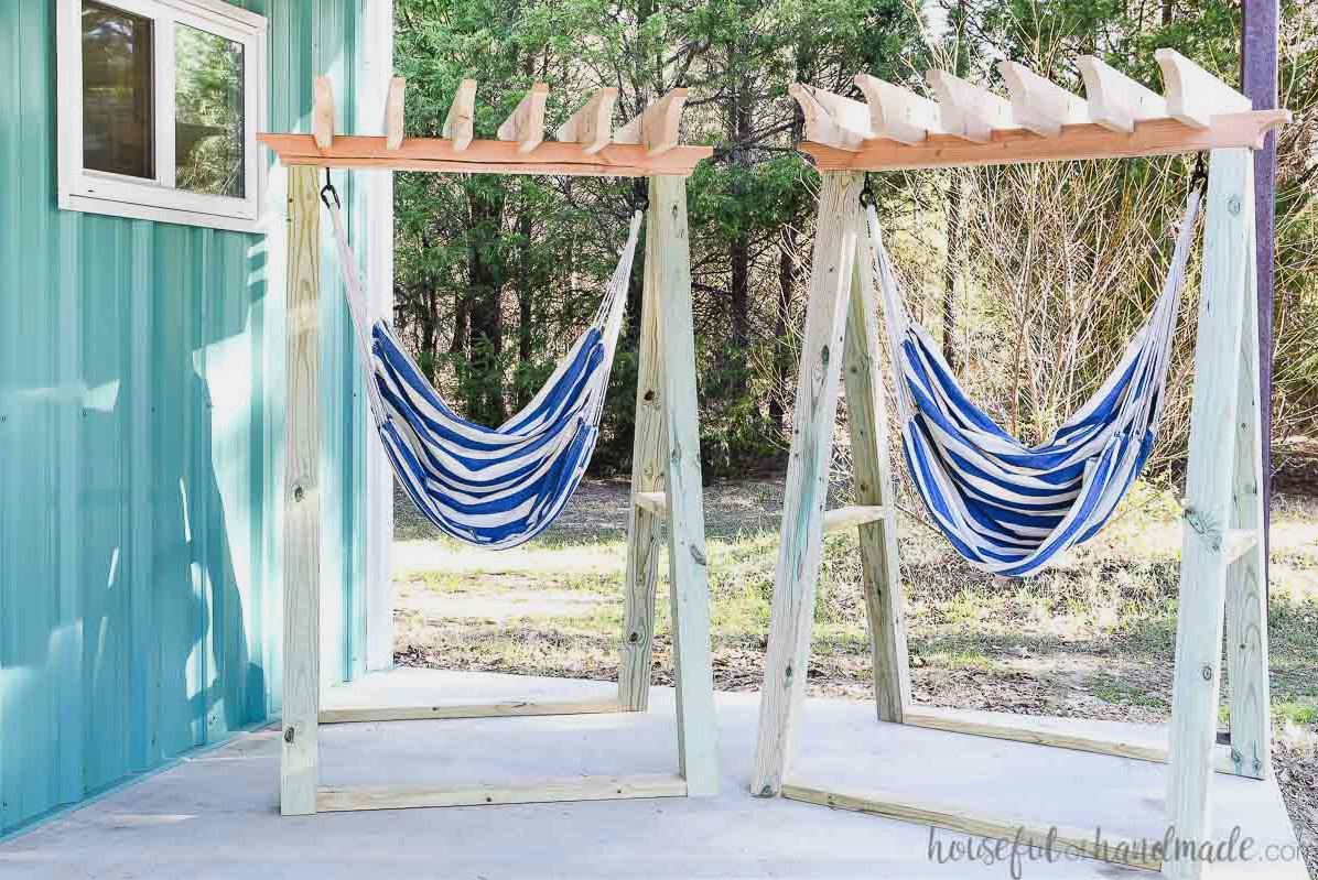 Two freestanding hammock chair stands placed on a porch beside an aqua metal building. 