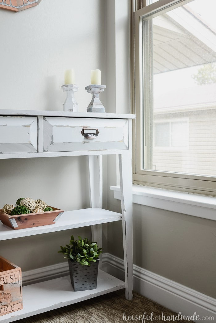 Create a lovely small console table for added storage and home decor. This DIY farmhouse console table features two spacious drawers and two shelves. An ideal storage solution for compact spaces. Housefulofhandmade.com White farmhouse console table positioned beside the window. This compact table includes drawers and two shelves. Housefulofhandmade.com